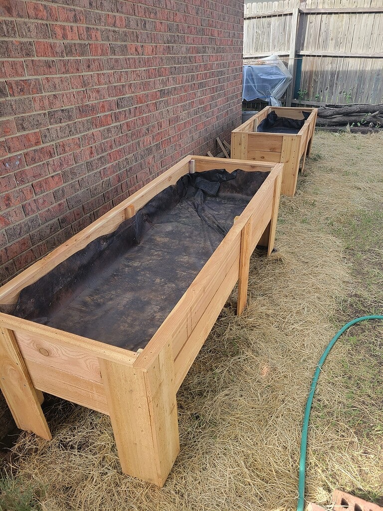 two handcrafted elevated wooden planter boxes installed side-by-side along a red brick house wall in a residential backyard
