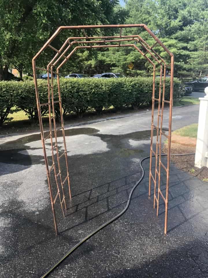 a freestanding metal garden arbor positioned on a sunlit asphalt driveway in a suburban neighborhood.
