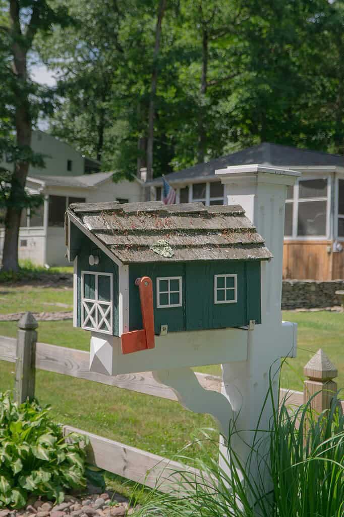 a whimsical, house-shaped mailbox mounted on a white wooden post and fence rail in a quiet residential setting.
