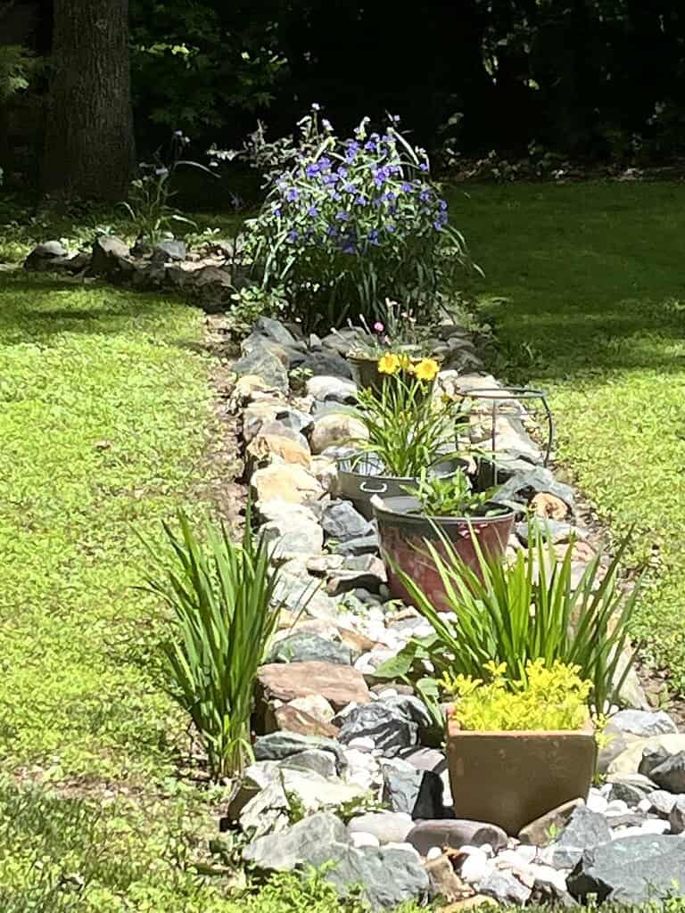 Narrow backyard rock garden border with natural river stones, colorful flowering plants, and decorative containers