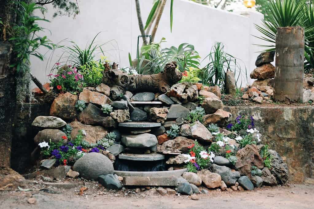 Layered backyard rock garden with stacked natural stones, small waterfall feature, succulents, and colorful flowers