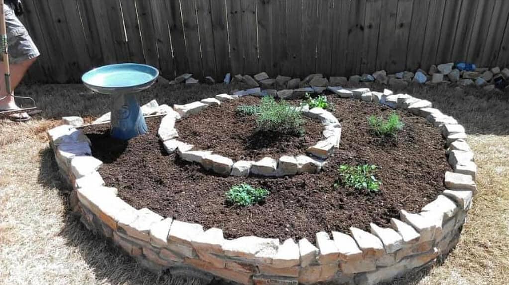Tiered circular rock garden with stacked stone edging, planted with drought-tolerant herbs like rosemary and lavender, plus a birdbath centerpiece