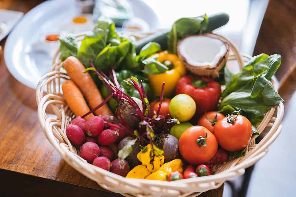 a wicker basket overflowing with freshly harvested organic vegetables arranged on a wooden table near a bright window