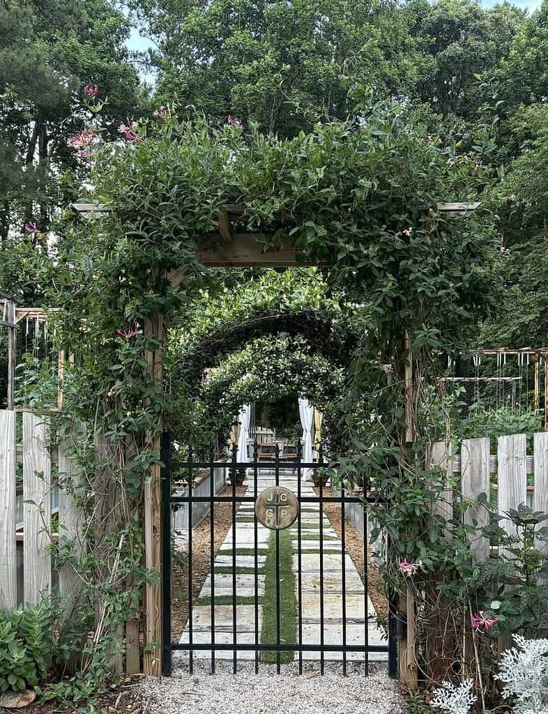 Garden entrance with vine-covered arbor, metal gate, and pathway leading to a backyard oasis
