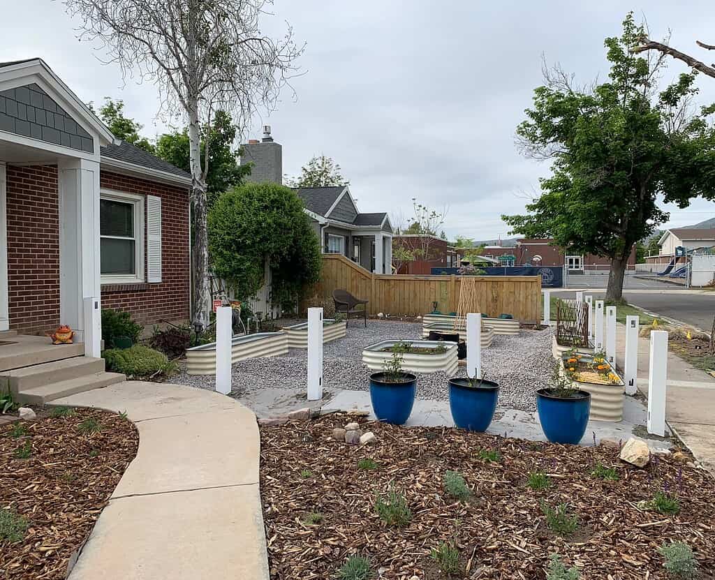 Front yard garden with raised metal beds, gravel paths, and blue planters near a brick house.