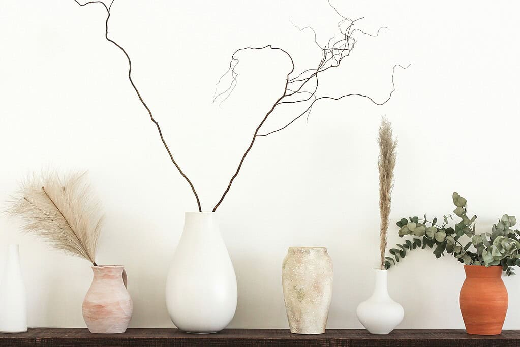Neutral-toned ceramic vases with dried branches, pampas grass, and greenery arranged on a wooden shelf against a minimalist white wall.