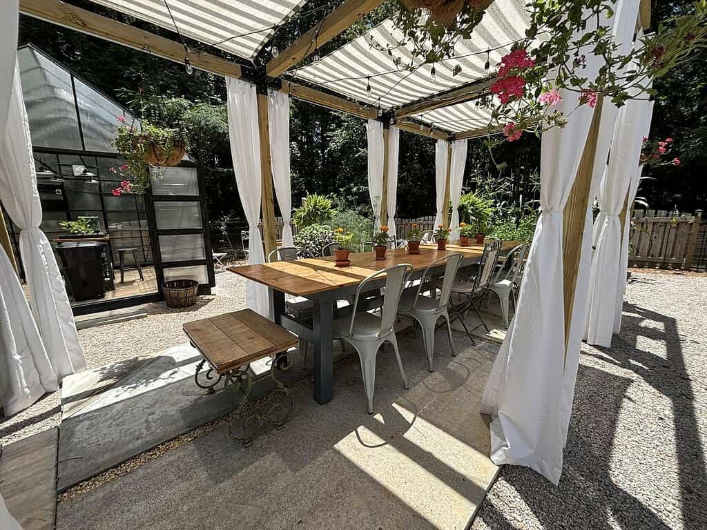 Outdoor pergola dining area with wooden table, white curtains, hanging plants, and cozy garden setting