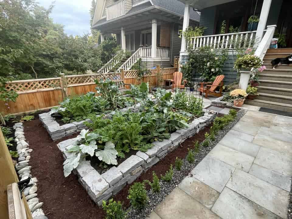 Front yard vegetable garden with raised stone beds beside a walkway and porch seating.
