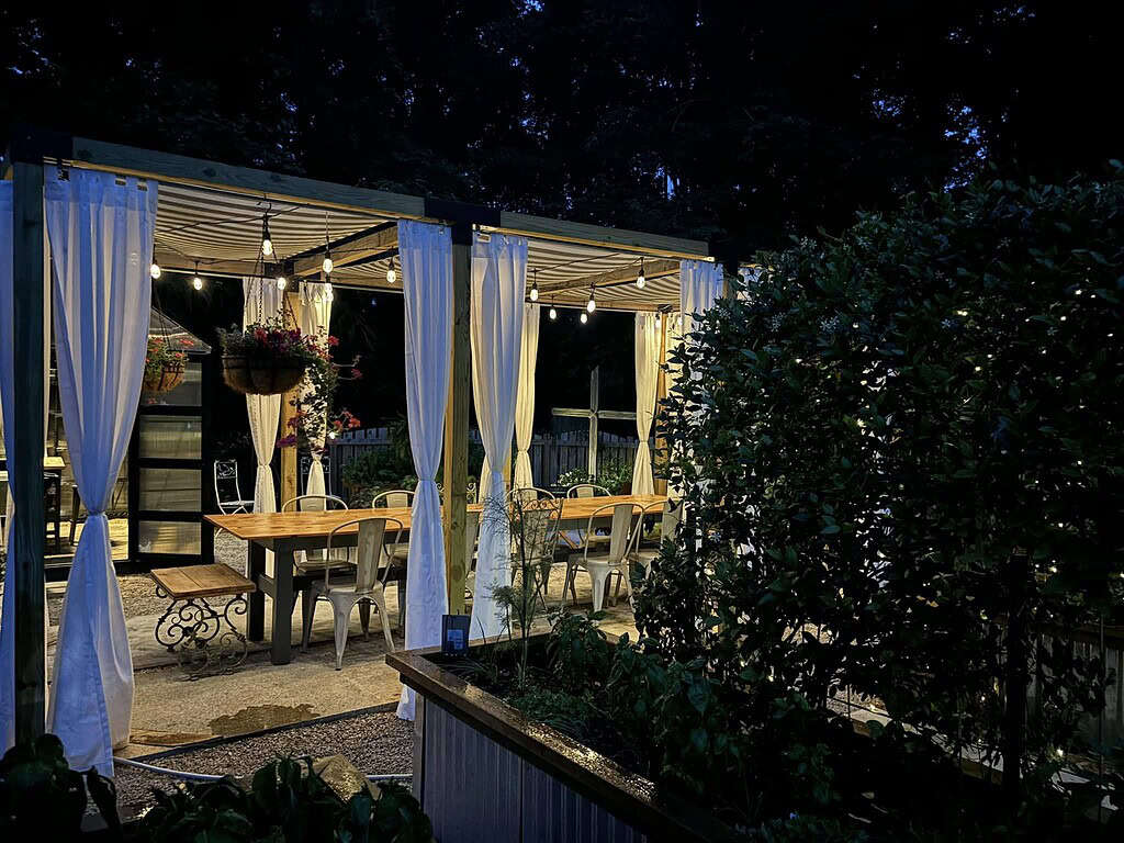 Backyard pergola dining area at night with string lights, white curtains, and cozy garden ambiance