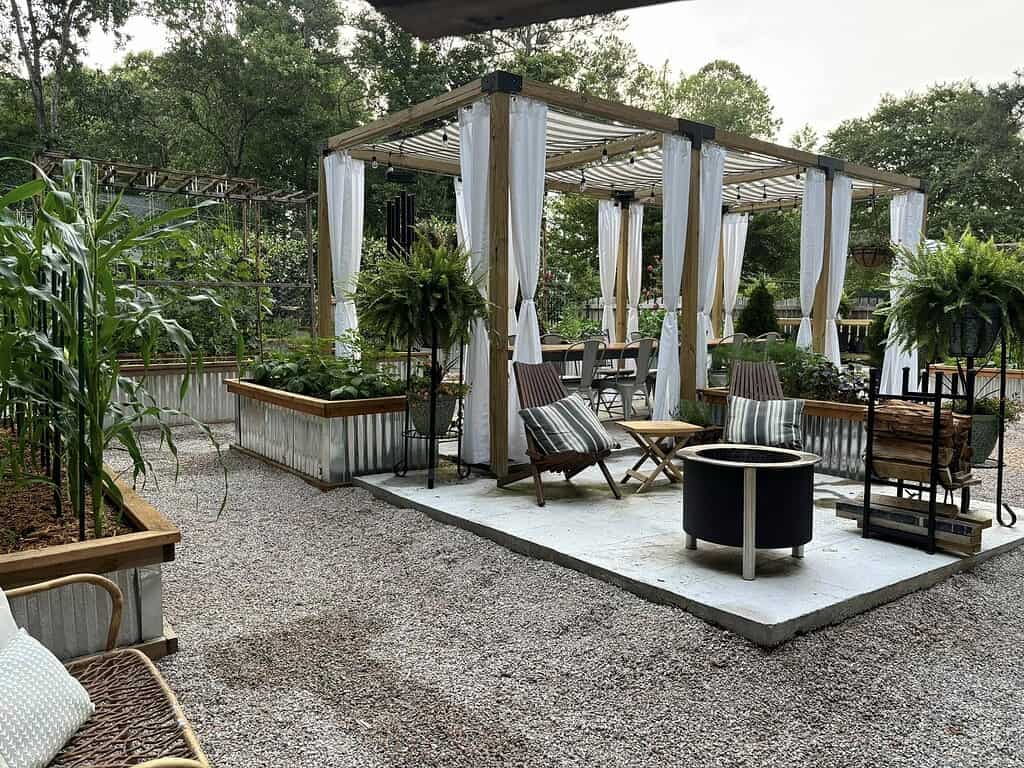 Backyard oasis with pergola seating area, fire pit, raised garden beds, and gravel patio surrounded by greenery
