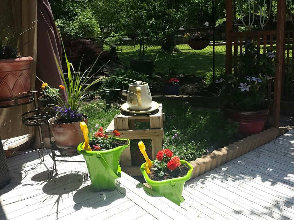 Backyard garden patio with potted flowers, watering cans, and shaded seating area creating a cozy outdoor space
