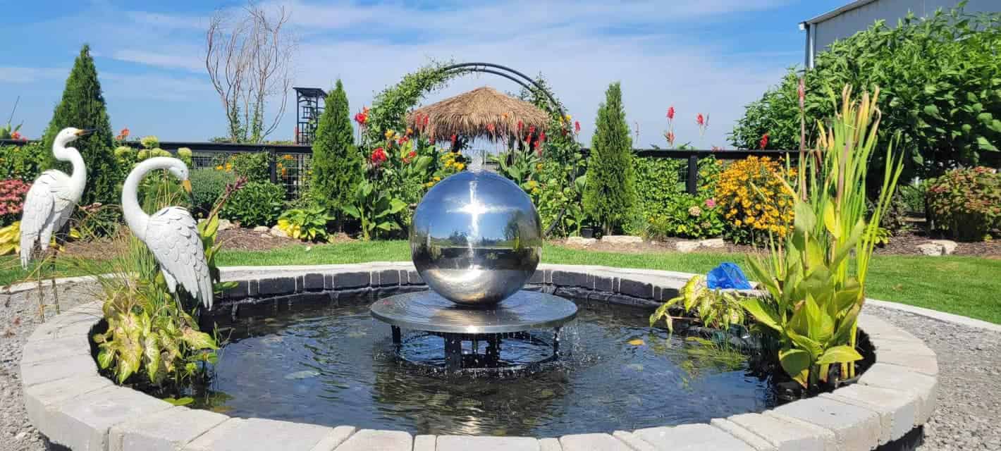 Outdoor water fountain with reflective metal sphere in a landscaped garden pond surrounded by flowers and decorative bird statues