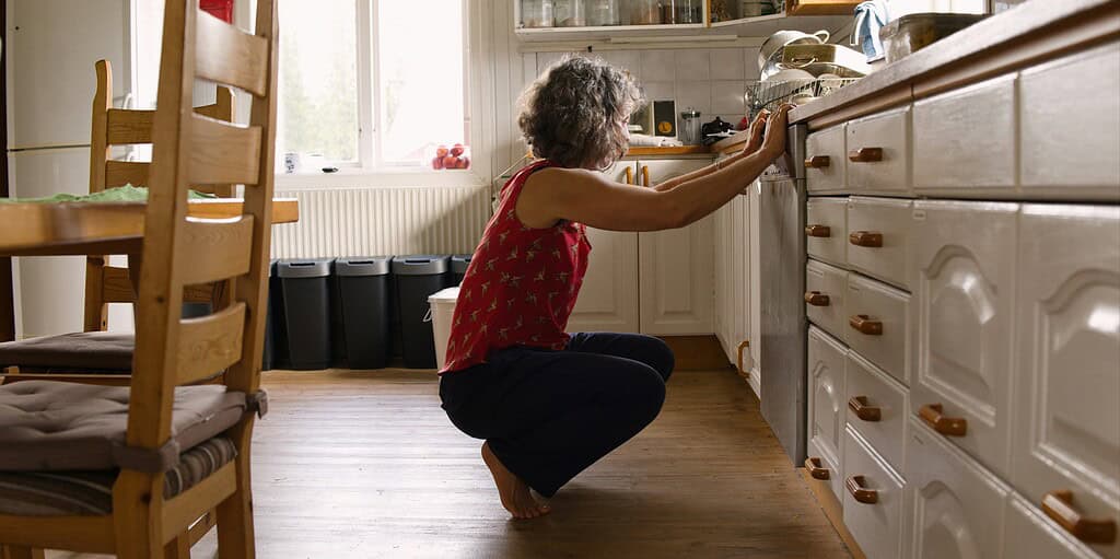 Person crouching in a kitchen, holding onto a cabinet for support.