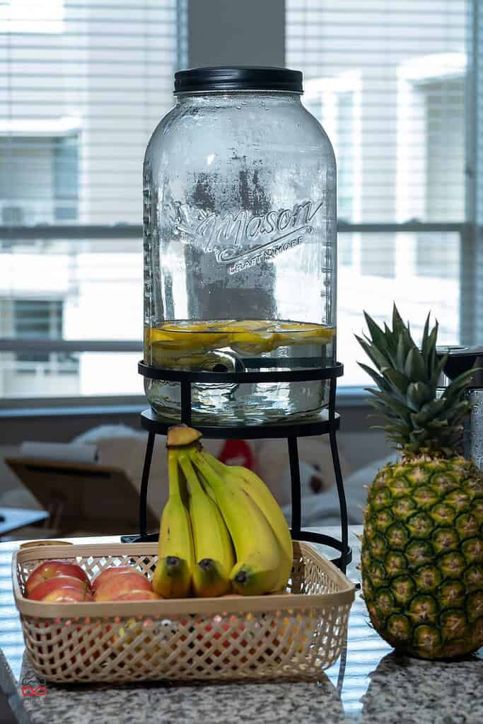 Large glass dispenser with fruit-infused water on a kitchen counter, with bananas, apples, and a pineapple nearby.