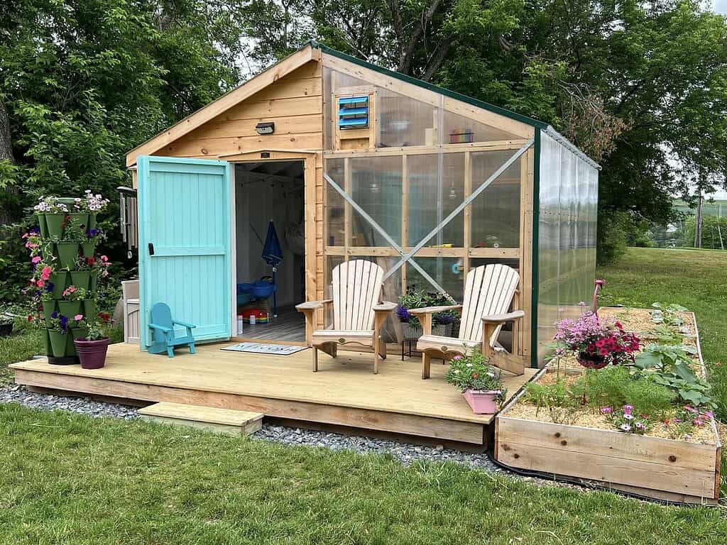 Small backyard greenhouse and shed with a wooden deck, turquoise door, two chairs, and surrounding raised garden beds and potted flowers