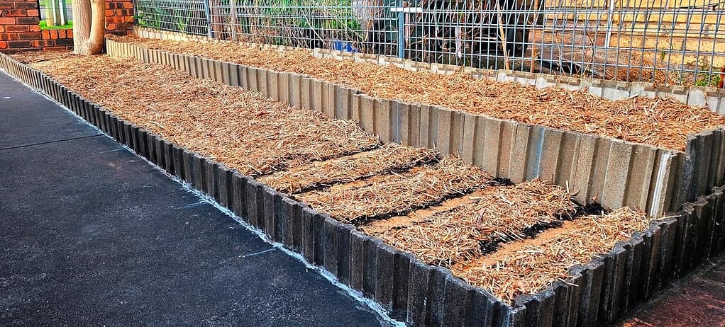 Long raised garden bed covered with mulch beside a sidewalk and fence.
