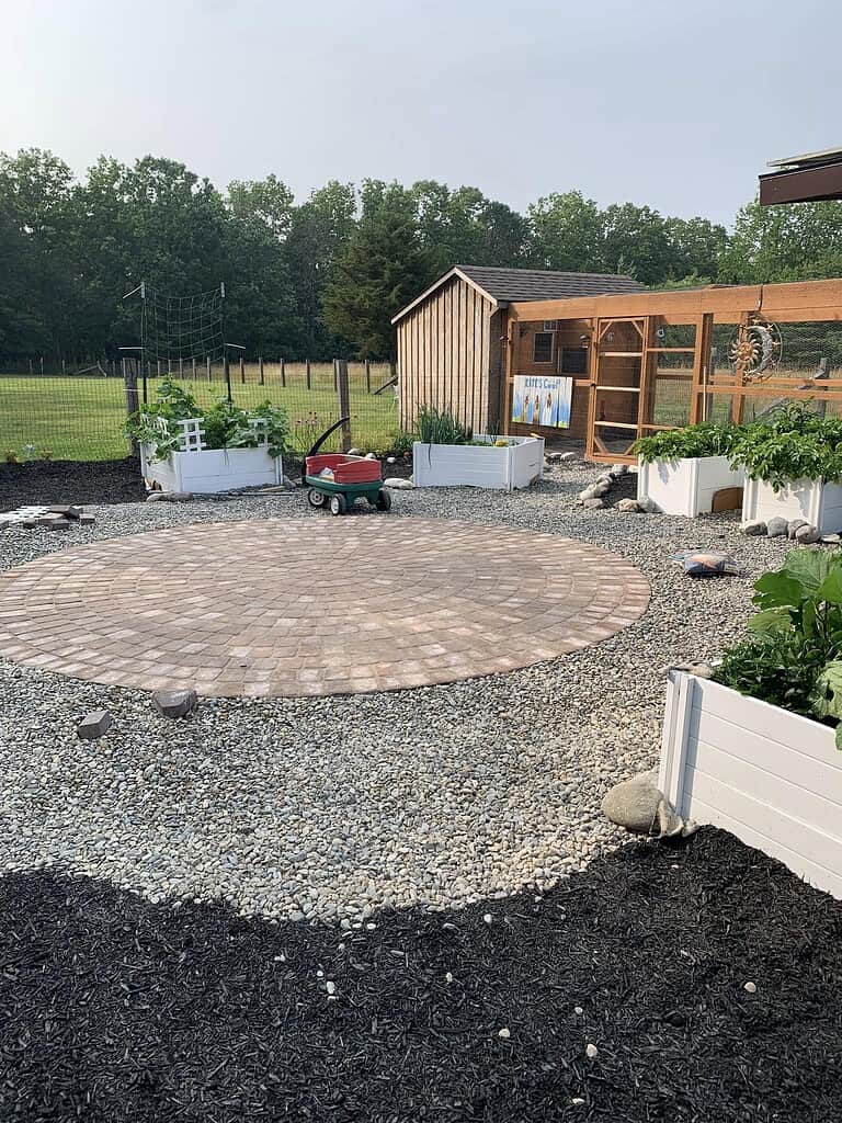 Large circular paver patio set within a gravel yard, with garden beds and a small wagon nearby.
