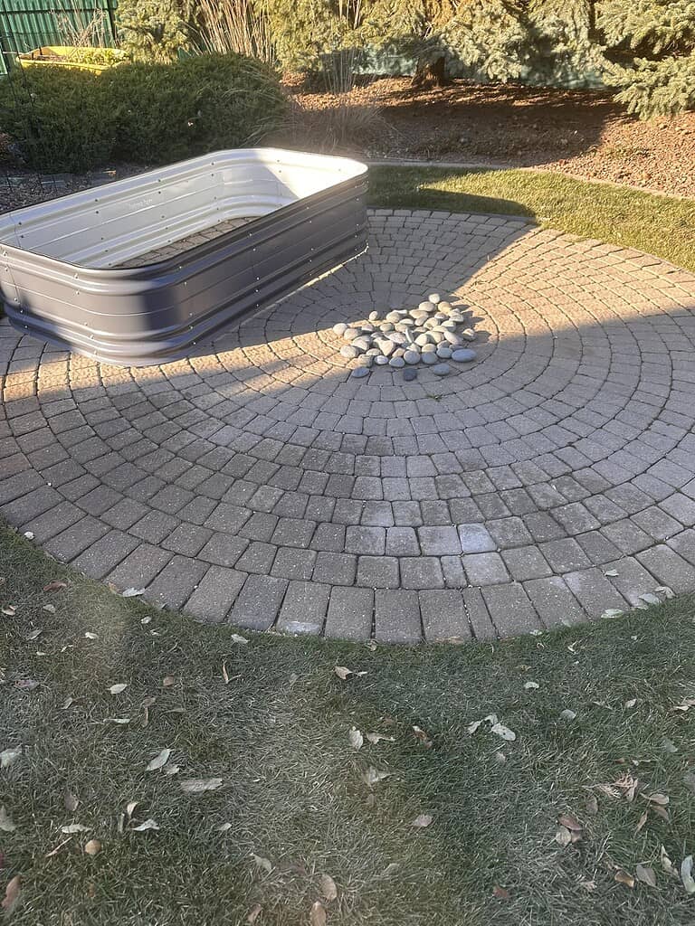 Circular stone paver patio with a central fire pit area and a metal stock tank nearby.
