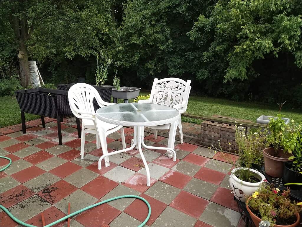 Checkered red and gray concrete paver patio with outdoor table, chairs, and garden items.