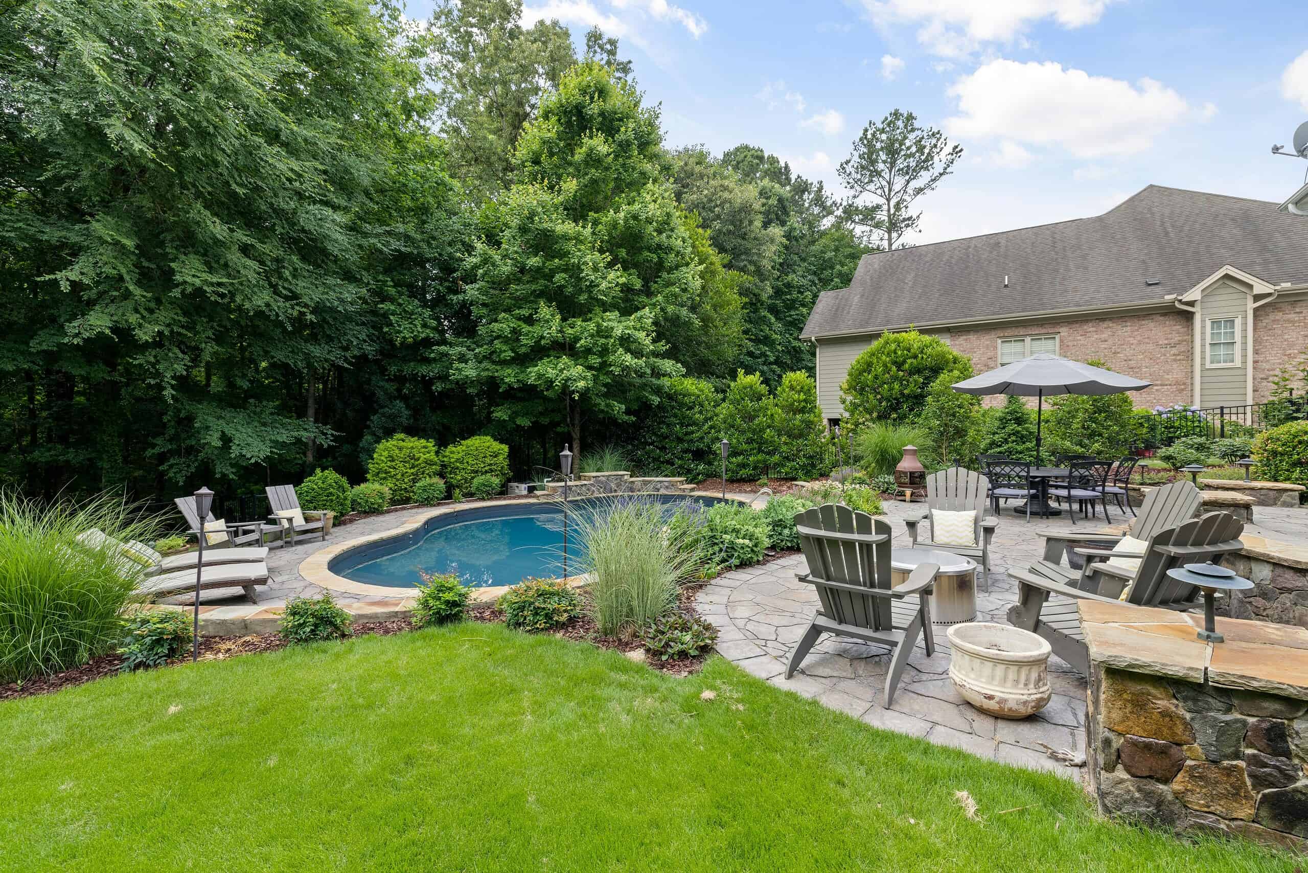 Backyard with a small curved pool, stone patio seating, dining set with umbrella, and lush trees surrounding a brick house.