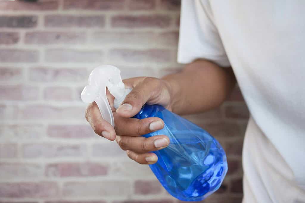Hand holding a blue spray bottle against a brick background.
