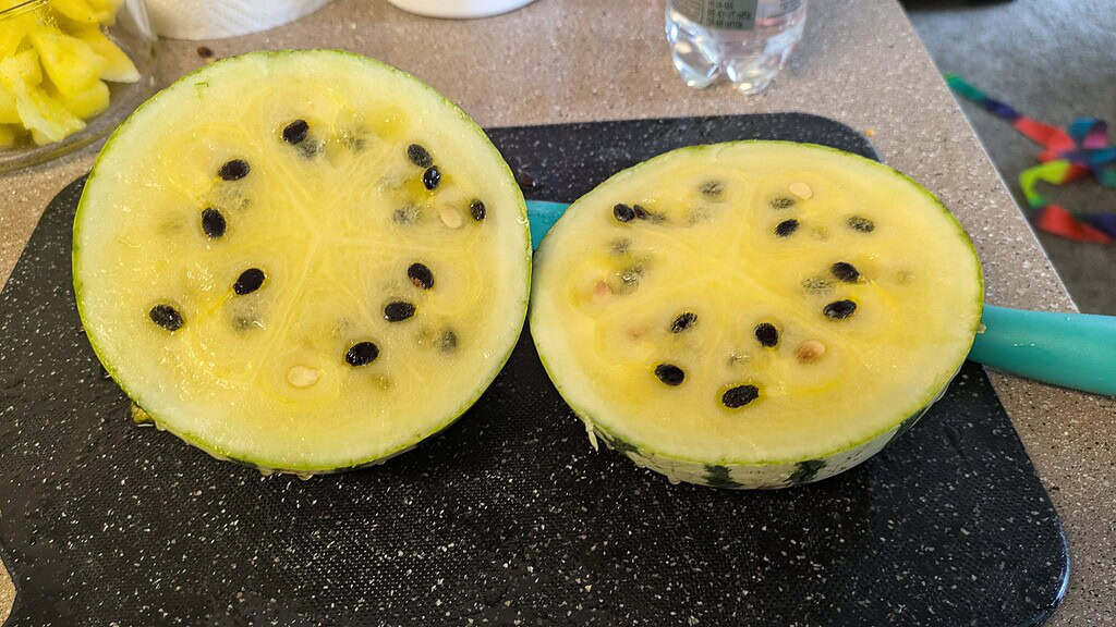Yellow-fleshed watermelon cut in half on a cutting board, showing black seeds and pale yellow interior.
