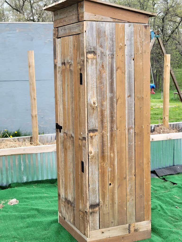 A tall narrow rustic wooden cabinet with vertical weathered planks, two black metal latch hinges, and a flat top, standing on artificial green grass in a backyard, with a raised garden bed, wooden fence posts, and a blue swing set visible in the background