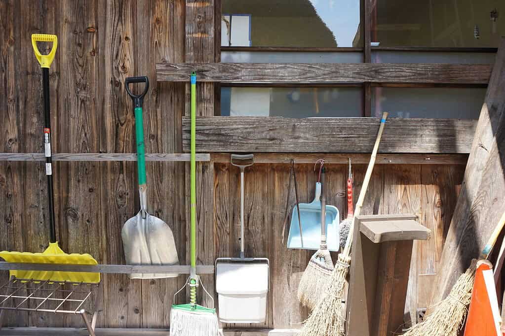 Several long-handled tools leaning against and hanging on a weathered wooden plank wall, including two shovels with yellow and green handles, a mop, a dustpan, a blue plastic scoop, a straw broom, and a wooden post, with a yellow snow pusher on the left and an orange traffic cone partially visible on the right