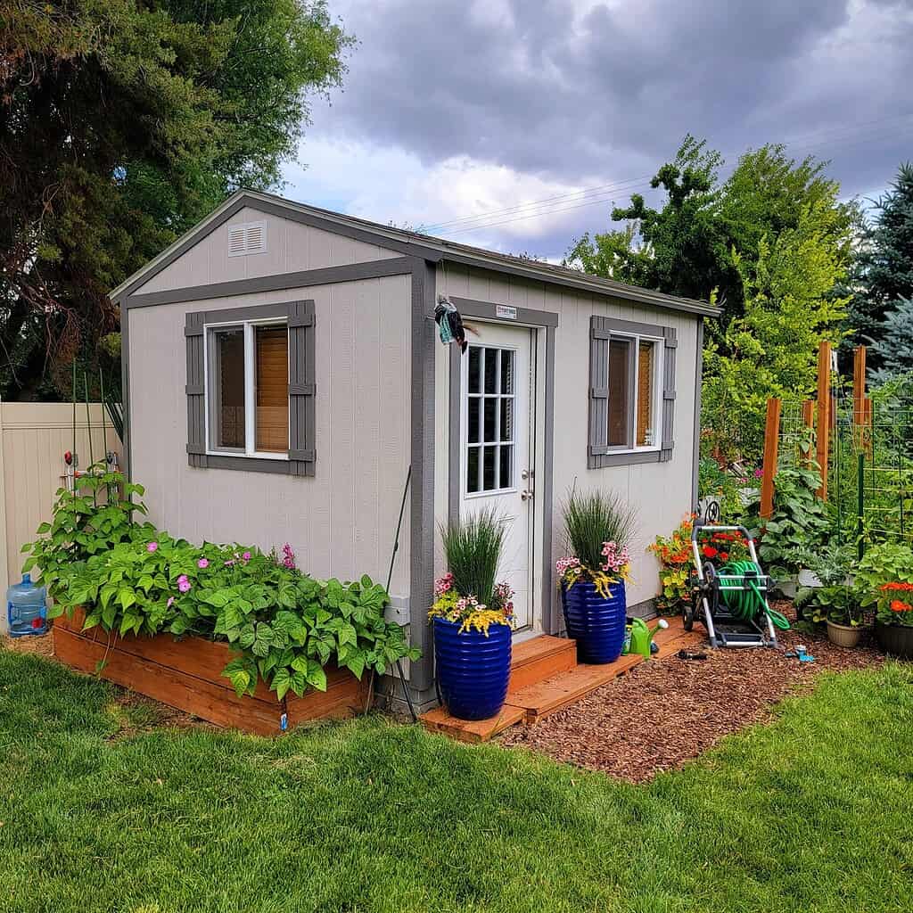 A light gray wooden storage shed with gray shutters, two windows, and a white door, sitting on a small wooden deck platform, surrounded by lush green plants, colorful flower pots, a raised garden bed, and a vegetable garden, with trees and a cloudy sky in the background