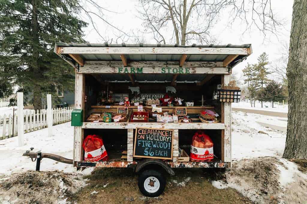 A small wooden farmstand with open shelves and a chalkboard sign, displaying red crates of produce, in a snow-covered yard with a white picket fence and bare trees.